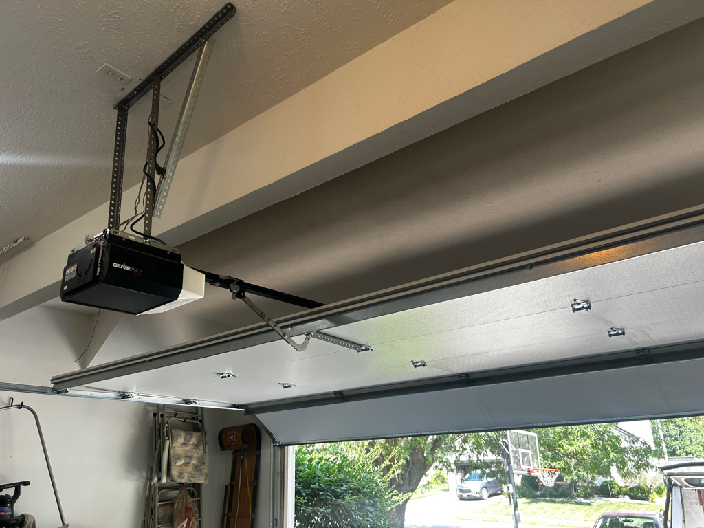 Open residential garage with overhead door, ceiling-mounted opener and rails, basketball hoop, lawn, trees, and sunny street outside.