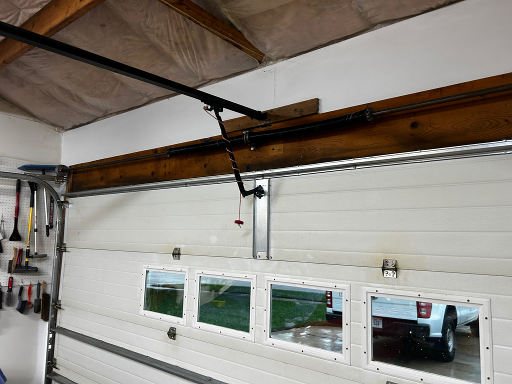 Interior of a residential garage featuring a white sectional door with four small windows, exposed wood beams, insulation, organized tools, and an opener rail ready for garage opener repair.