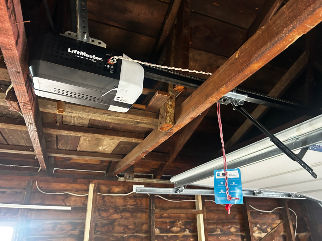 A LiftMaster garage door opener hangs from exposed wooden ceiling beams, with a red emergency-release cord and blue instruction tag; rustic, utilitarian interior.