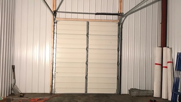 A closed white sectional garage door fills a metal-sided workshop, with overhead tracks and rolled materials leaning at right; the scene feels utilitarian.